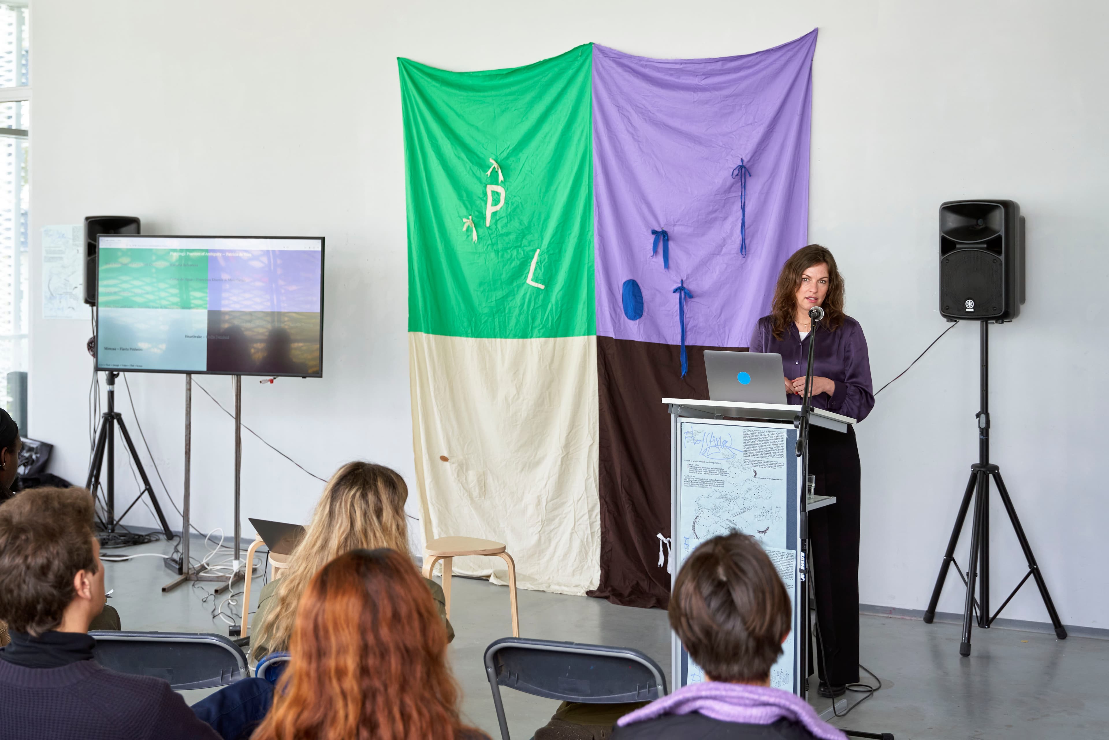 The image shows a woman standing in front of a large piece fabric divided into four colours: green, lilac, cream, and brown. A screen next to it depicts a similar colour scheme. The audience sits in the foreground.   