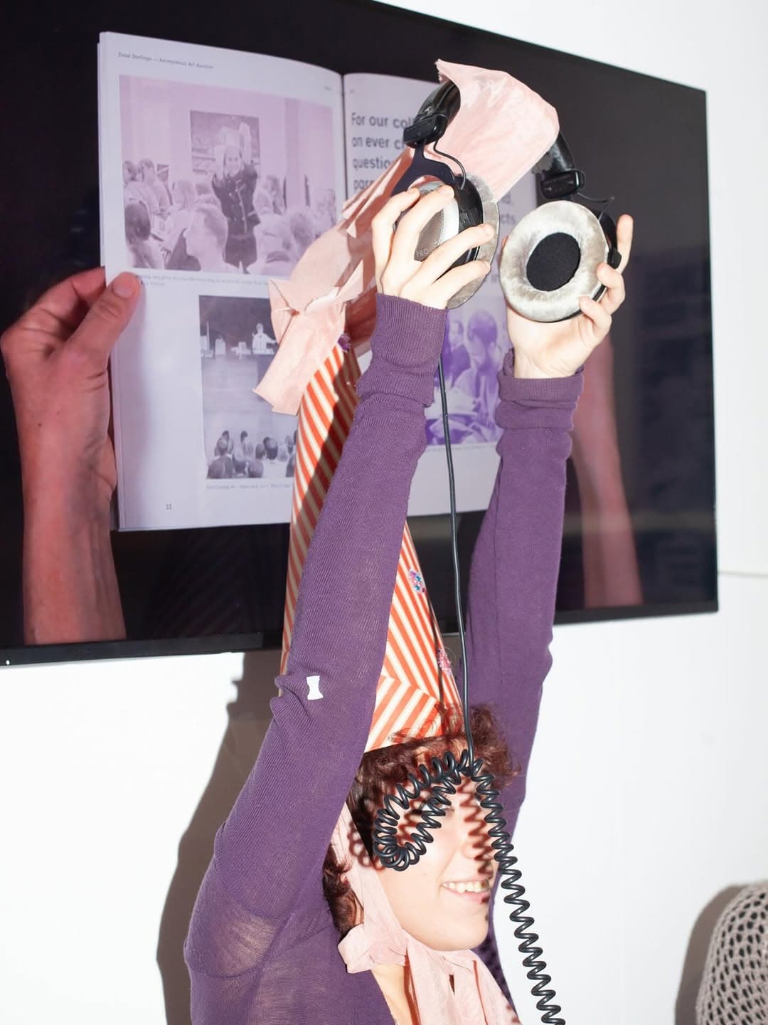 The image shows a person wearing long sleeve purple vest and wearing a tall striped orange party hat. She is holding a set of headphones above her head. 