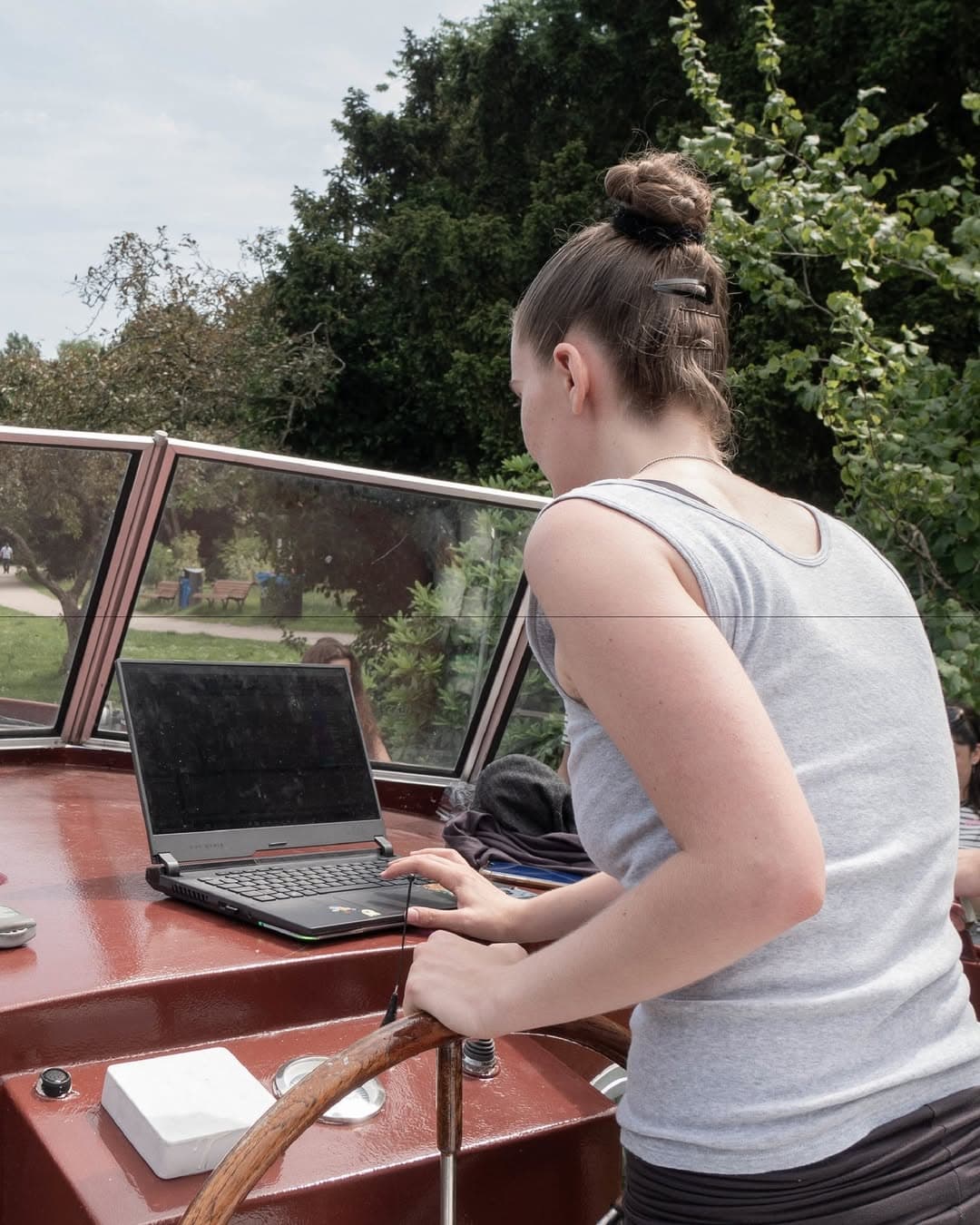 The image shows someone at the helm of a boat while looking at a laptop. It is summer. There are trees in the background.  