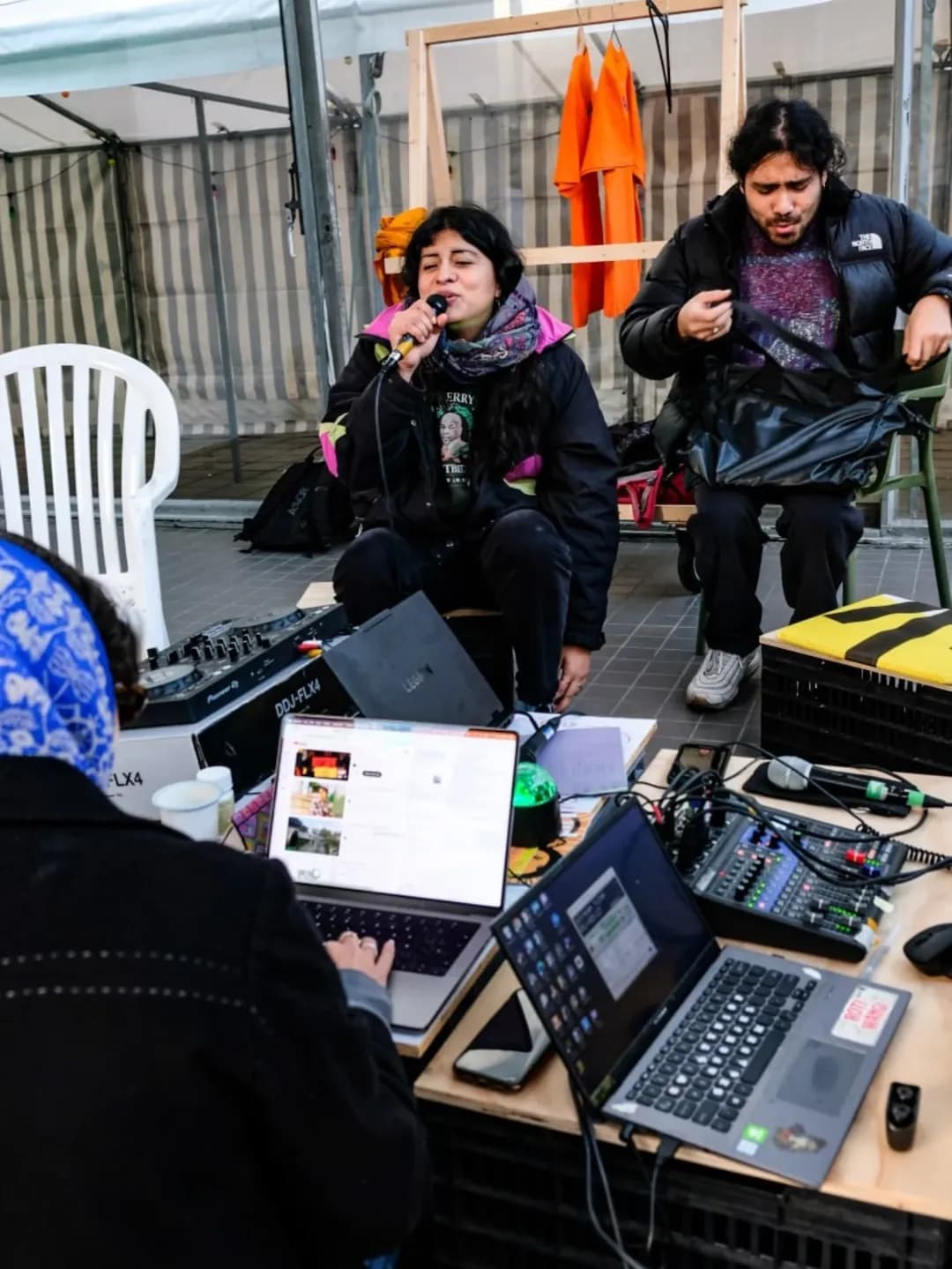 The image shows a radio show livestream setup. There are laptops and equipment in the foreground. One person is speaking into a microphone. They are wearing dark jackets and they are outside.