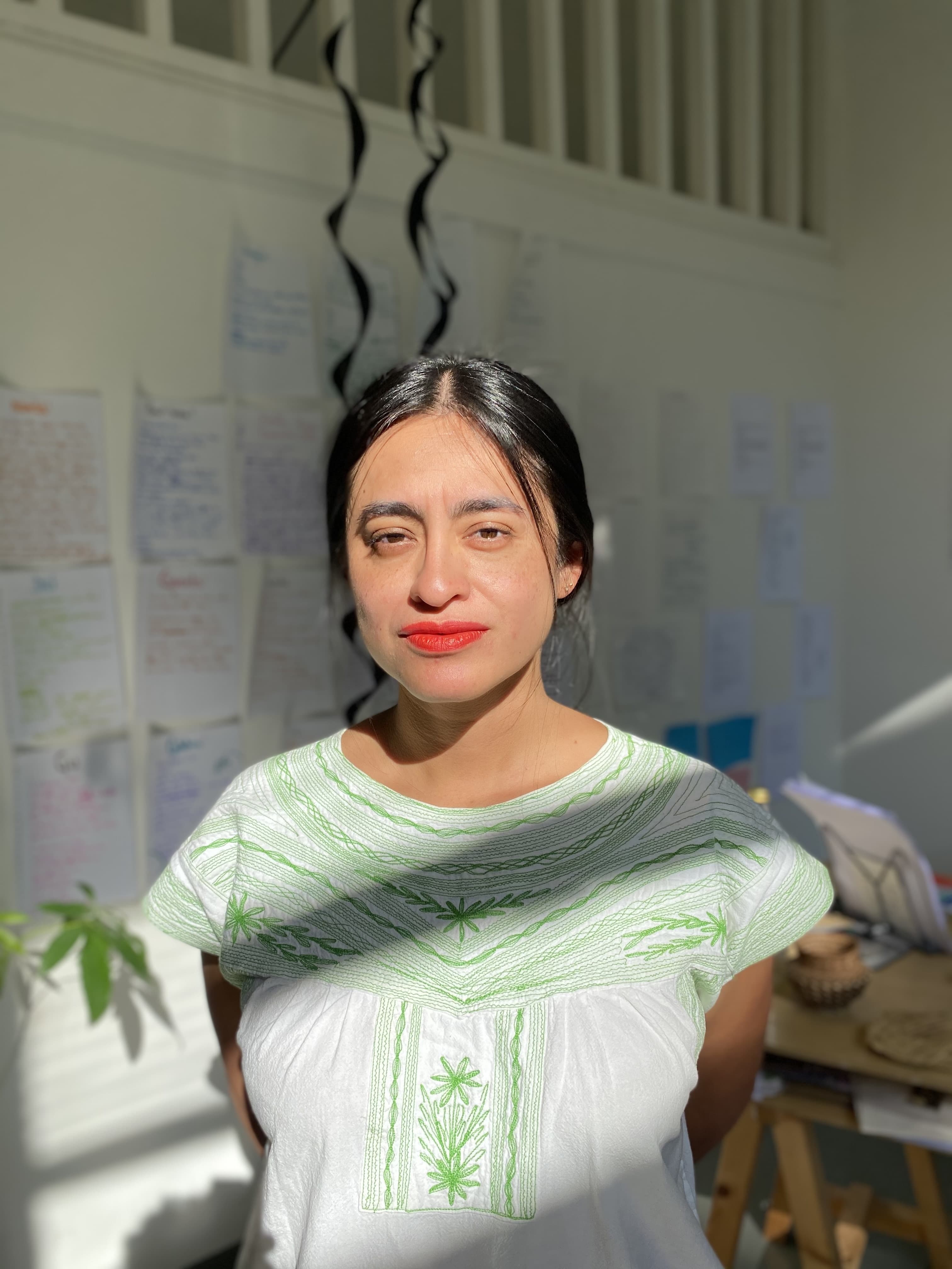 Eva Posas stands in a sunlit room wearing a white blouse with green embroidery. Her arms are crossed behind her back, and she smiles gently at the camera with red lipstick. Behind her, the wall is covered with handwritten notes, papers, and two black ribbons hanging from above. 