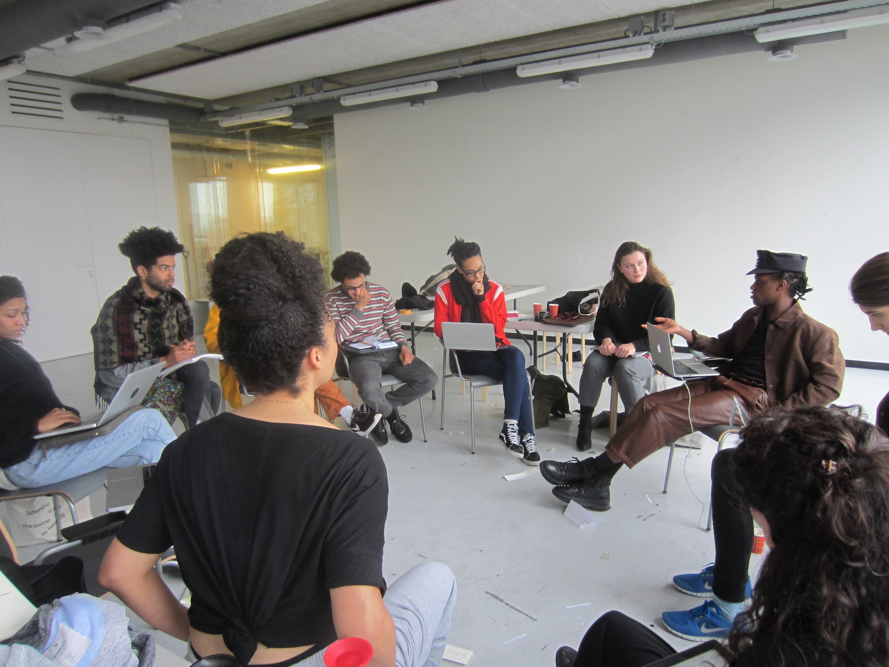 The image shows a group of people sitting in a circle on laptops in a workshop setting.